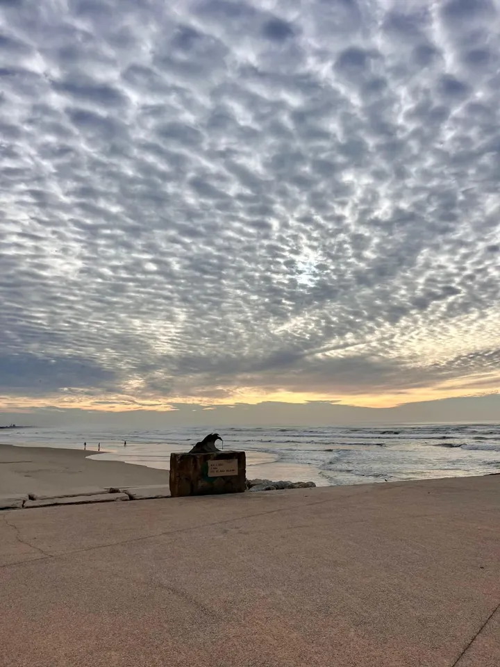 The beach at Cabedelo in late afternoon light, a rippled mackerel sky over a wide empty beach, with a small stone monument on the promenade in the foreground.